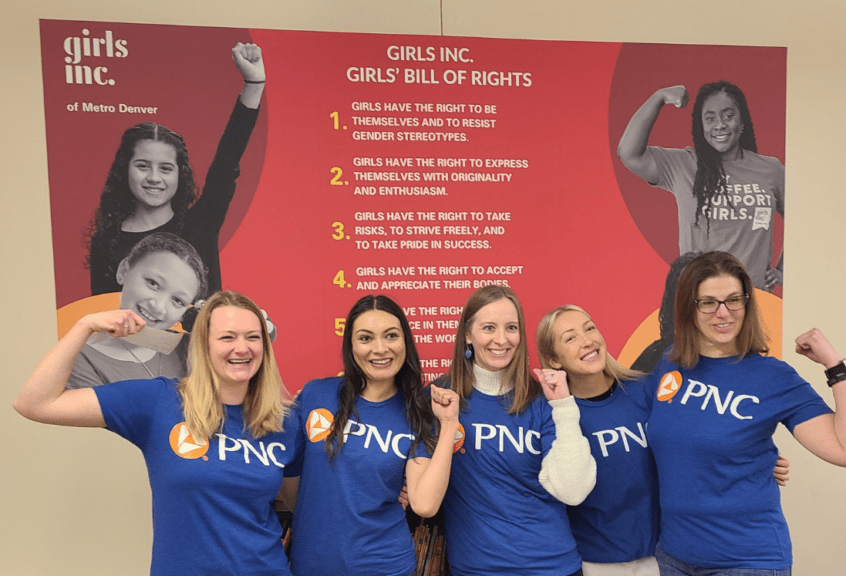 Group of five adult women volunteers smiling with arms raised in front of a sign of Girls Inc.'s Girls' Bill of Rights